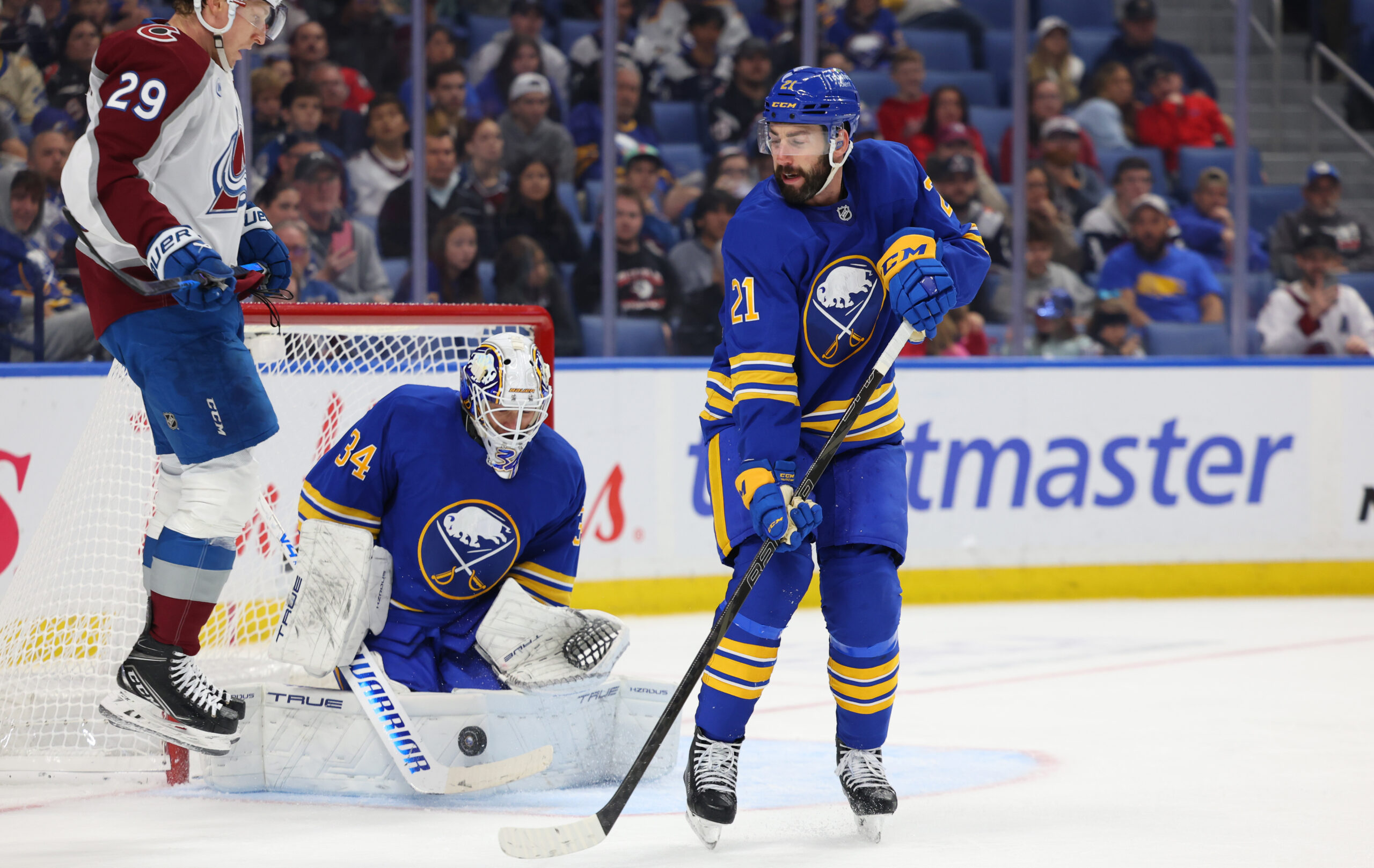 Oct 13, 2025; Buffalo, New York, USA;  Buffalo Sabres goaltender Alex Lyon (34) makes a save as Colorado Avalanche center Nathan MacKinnon (29) and Buffalo Sabres defenseman Conor Timmins (21) wait for a rebound during the third period at KeyBank Center. Mandatory Credit: Timothy T. Ludwig-Imagn Images